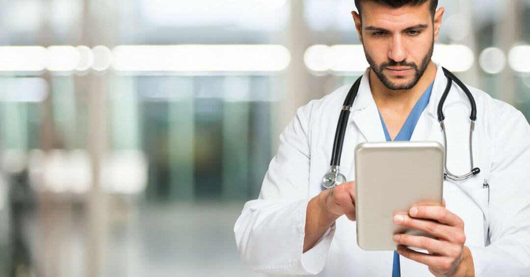A doctor in a lab coat is intently looking at a tablet in a clinical setting