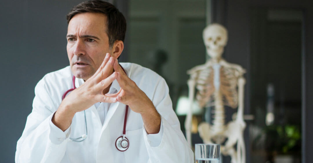 A pensive doctor with a stethoscope is seated next to a skeleton model in an office