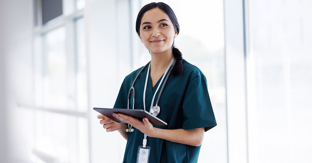 A cheerful female nurse in scrubs is holding a clipboard in a bright, windowed room