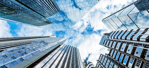 Low-angle wide shot looking straight up at glass-and-steel skyscrapers against a cloudy blue sky.   