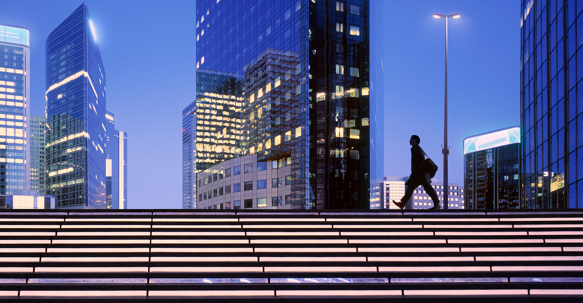 A silhouetted person walks across illuminated steps amid glowing glass skyscrapers in a modern city at dusk.