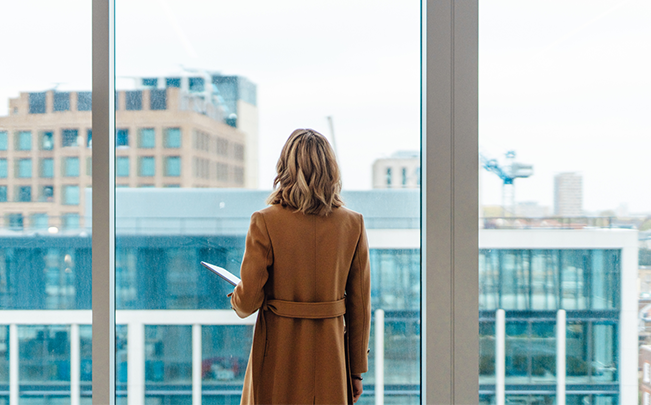 Businesswoman looking out over city buildings from a modern high-rise office window.
