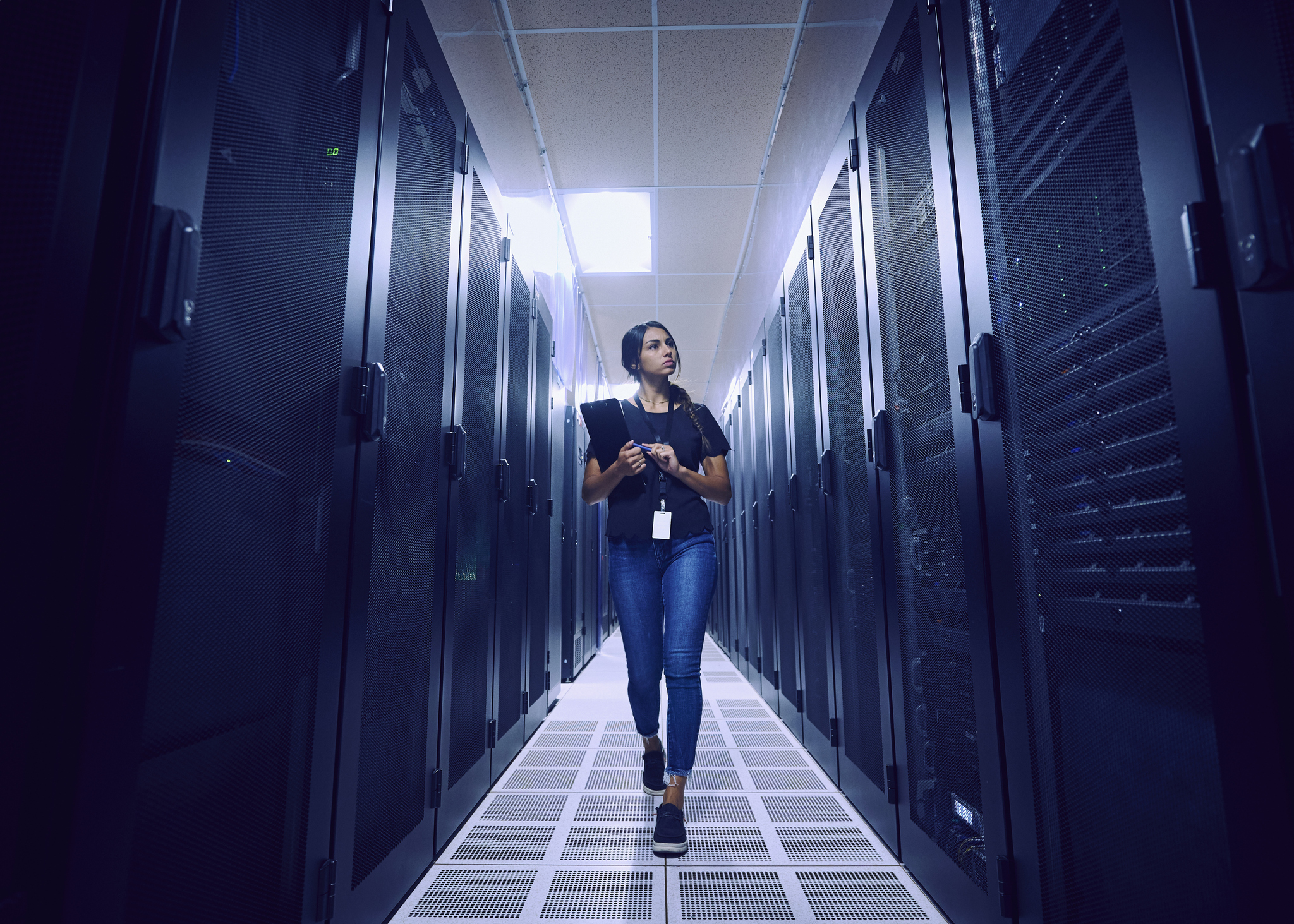 A woman walks through a server room aisle lined with tall data racks, holding a tablet and wearing an ID badge.