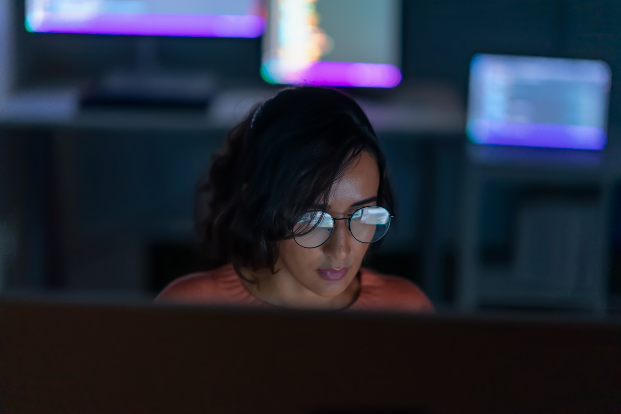 A person wearing glasses focuses intently on a computer screen in a dimly lit workspace with glowing monitors in the background.