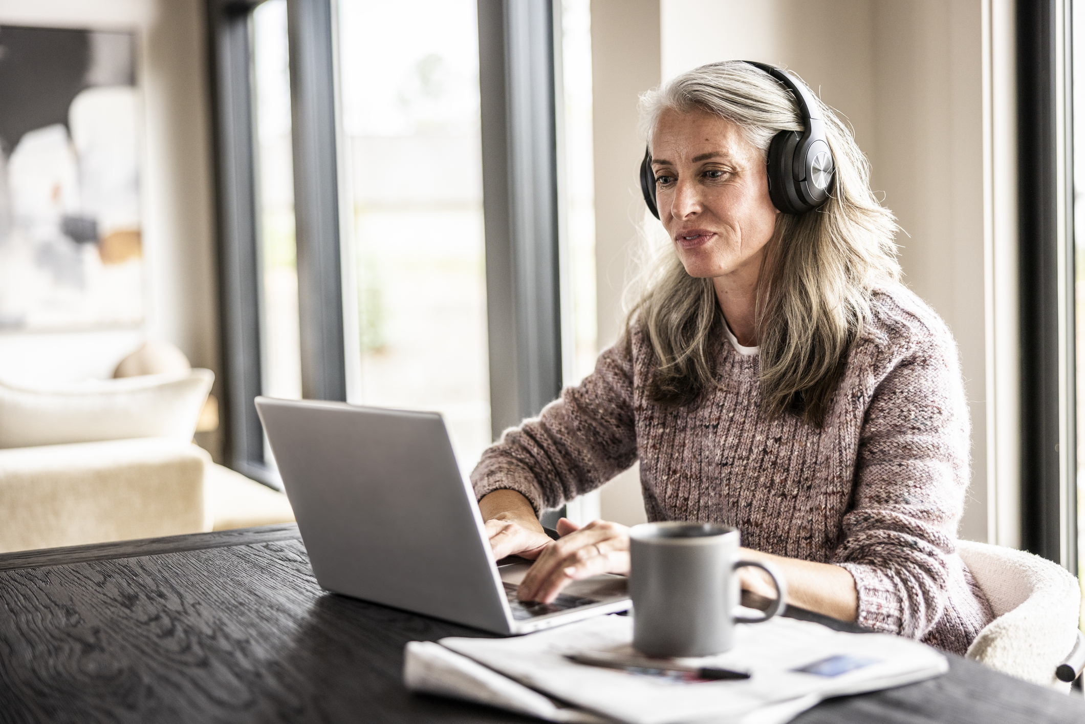 A gray-haired woman wearing headphones works on her laptop at a wooden table, a coffee mug nearby, in a softly lit home setting.