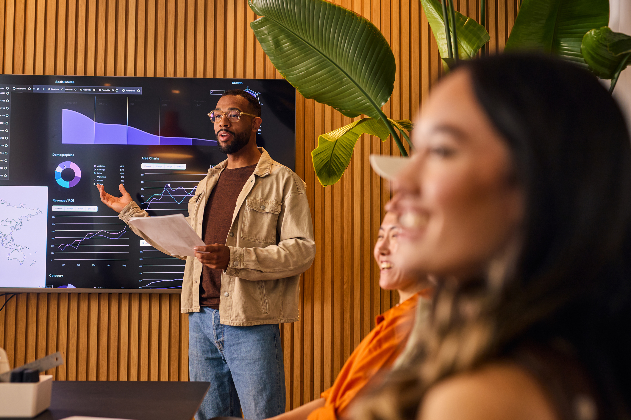 A man presents data on a screen in a modern office while colleagues listen in the foreground. 📊