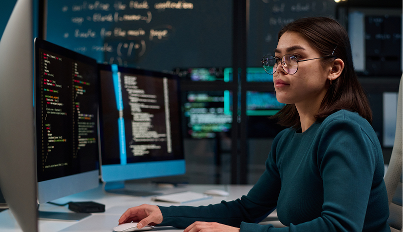 Female software developer working on code at a multi-monitor workstation in a modern office.
