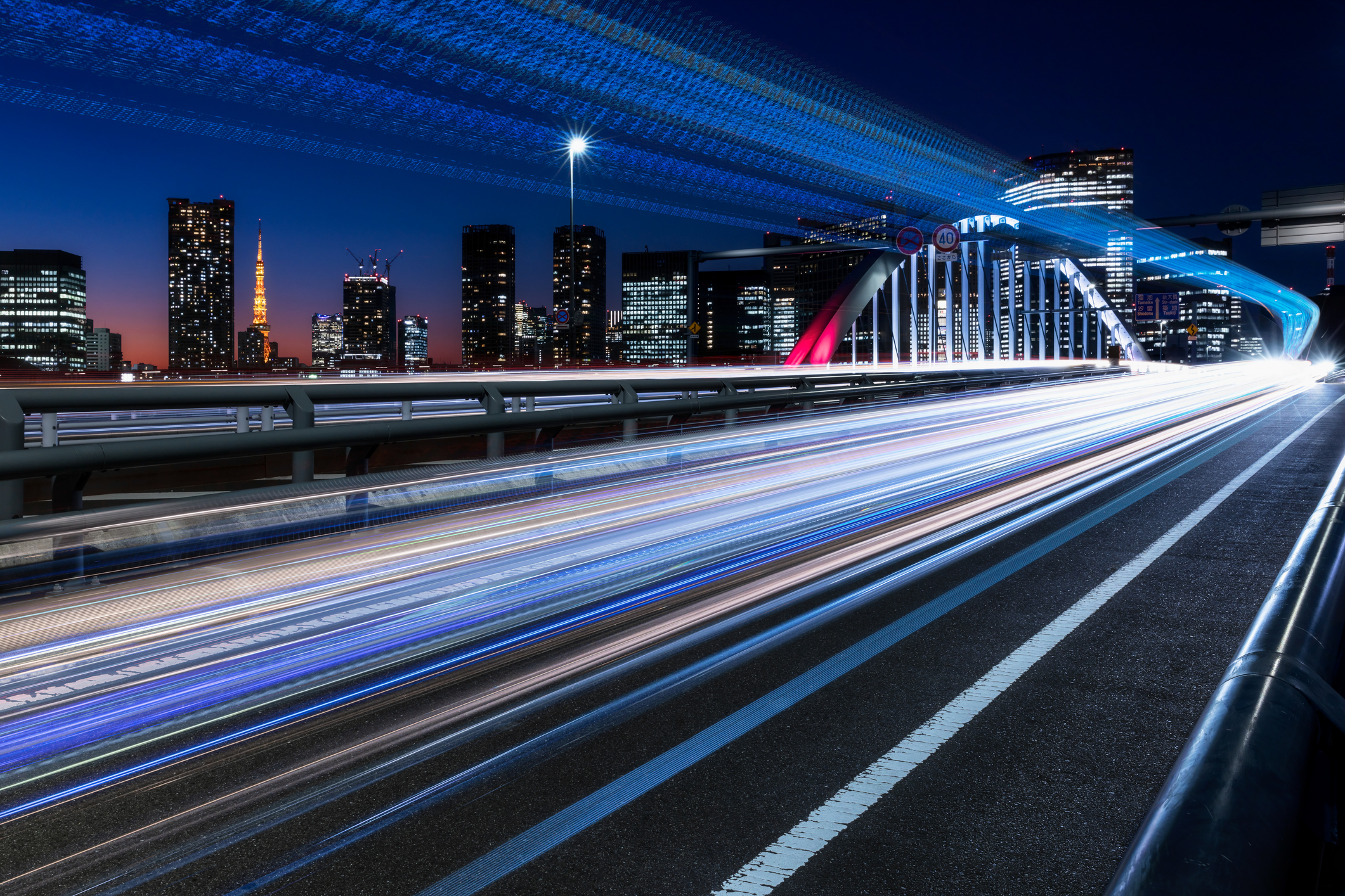 Long-exposure light trails streak across a city bridge at night, with illuminated skyscrapers glowing against the dark sky. 
