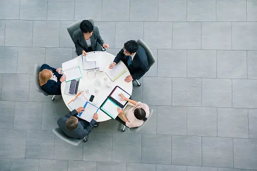 Five people in business attire seated around a circular table with laptops and documents