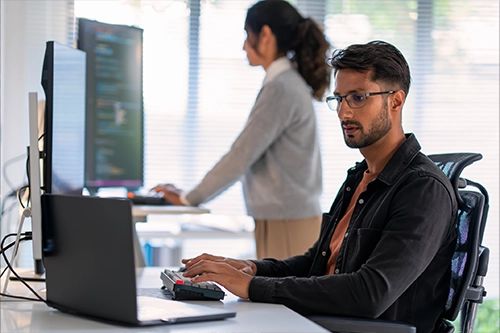 A man works at a computer in a modern office while a colleague stands at a desk in the background.