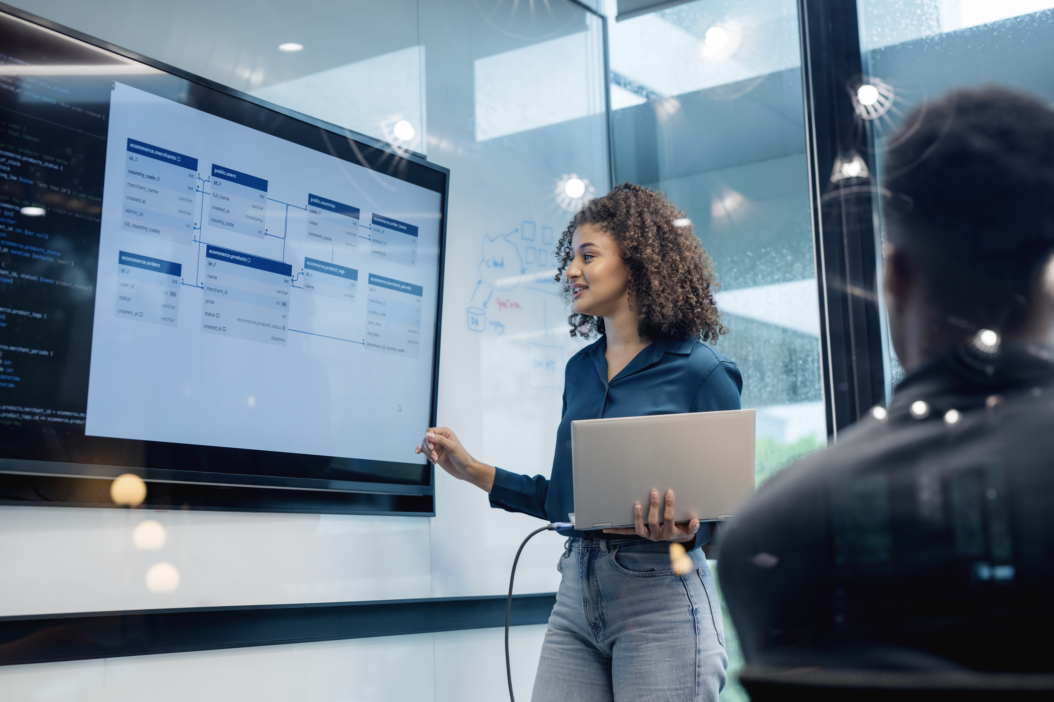 A woman holds a laptop while presenting information on a large screen during a meeting. 