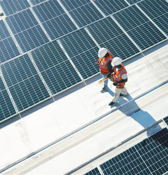 Two workers in helmets and vests walking among large solar panels on a rooftop.