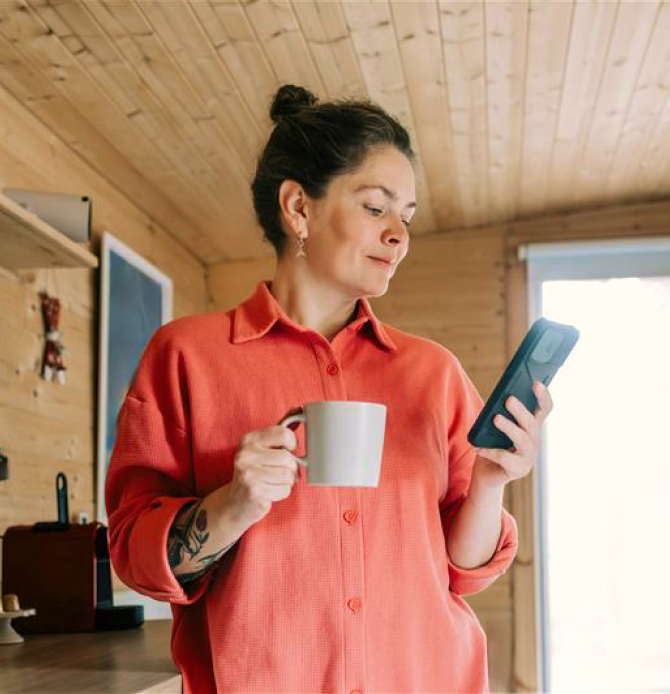 A person in a coral shirt holding a mug and looking at a smartphone indoors