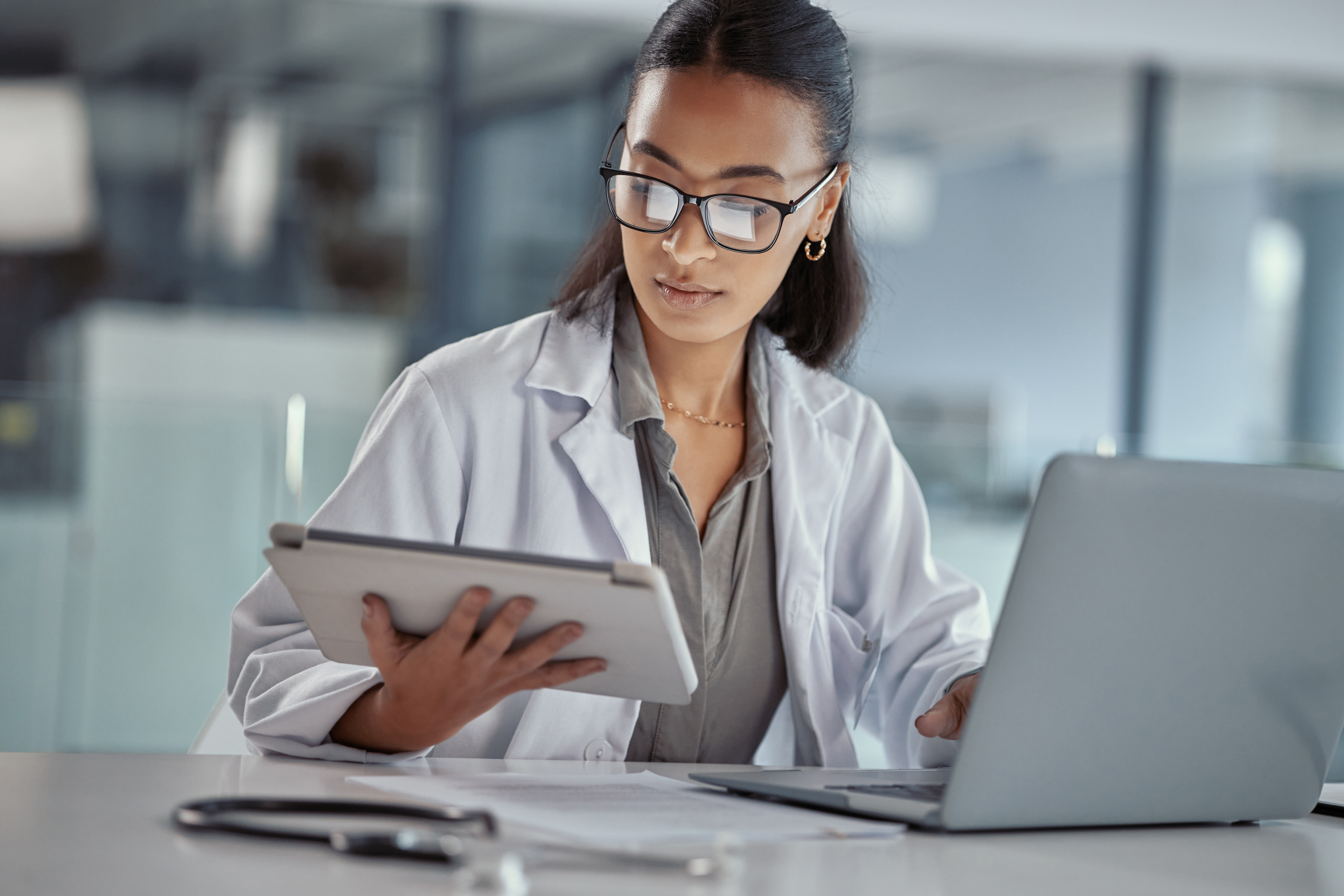 Doctor reviewing patient records on a tablet while working at a laptop in a modern medical office.