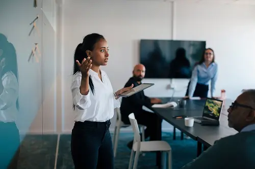 A woman presents to colleagues in a modern office setting with a table, chairs, and a wall-mounted screen