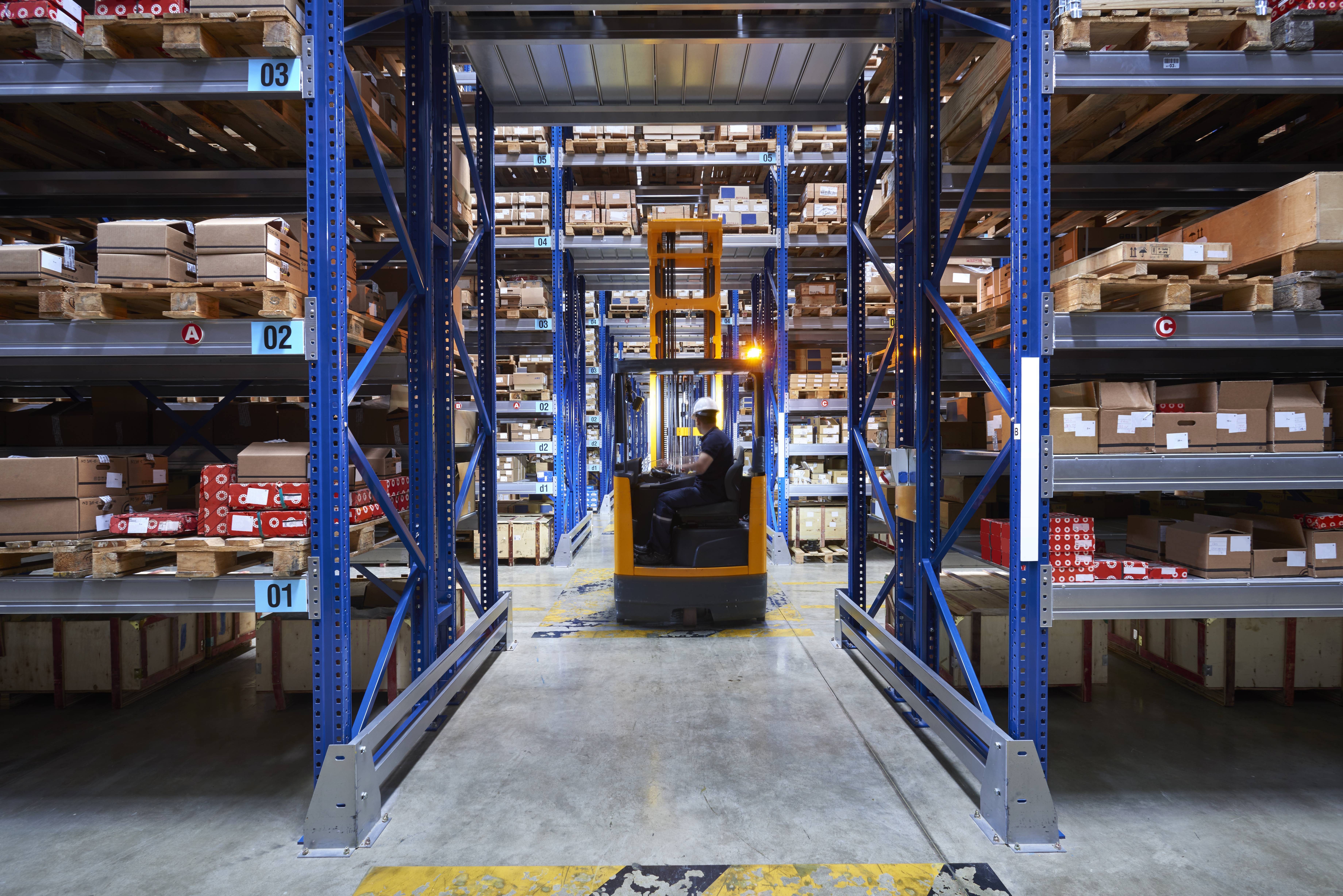 Yellow forklift driving down a narrow aisle lined with tall blue storage racks in a warehouse.   