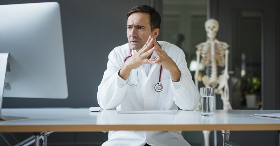 A male doctor in a white coat sitting at a desk with his hands steepled, looking focused at a computer screen in a medical office with a skeleton model in the background.